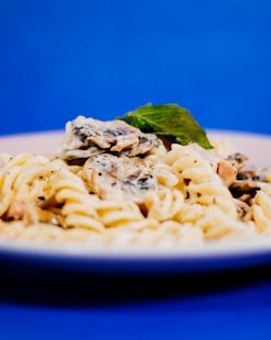 A close-up view of a plate of creamy pasta with mushrooms and a garnish of fresh basil set against a vibrant blue background. The dish appears to be rich and appetizing, showcasing a texture that suggests it has been freshly prepared.