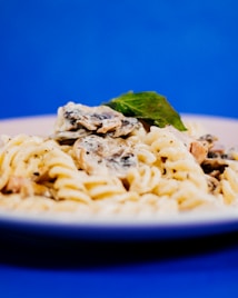 A close-up view of a plate of creamy pasta with mushrooms and a garnish of fresh basil set against a vibrant blue background. The dish appears to be rich and appetizing, showcasing a texture that suggests it has been freshly prepared.