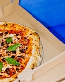 A freshly baked pizza with a golden-brown crust, topped with vibrant red sauce, melted cheese, scattered basil leaves, and slightly charred spots. The pizza rests inside an open cardboard delivery box against a bright blue background.