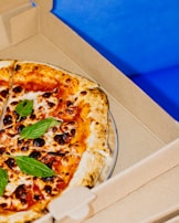 A freshly baked pizza with a golden-brown crust, topped with vibrant red sauce, melted cheese, scattered basil leaves, and slightly charred spots. The pizza rests inside an open cardboard delivery box against a bright blue background.