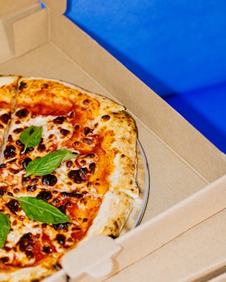 A freshly baked pizza with a golden-brown crust, topped with vibrant red sauce, melted cheese, scattered basil leaves, and slightly charred spots. The pizza rests inside an open cardboard delivery box against a bright blue background.