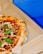 A freshly baked pizza with a golden-brown crust, topped with vibrant red sauce, melted cheese, scattered basil leaves, and slightly charred spots. The pizza rests inside an open cardboard delivery box against a bright blue background.