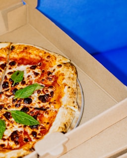 A freshly baked pizza with a golden-brown crust, topped with vibrant red sauce, melted cheese, scattered basil leaves, and slightly charred spots. The pizza rests inside an open cardboard delivery box against a bright blue background.