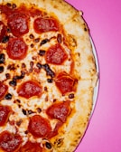 A delicious pepperoni pizza with golden-brown crust and melted cheese is positioned on a vibrant pink background. The pepperoni slices are evenly distributed across the pizza surface, and the cheese is slightly browned in some areas, adding texture.