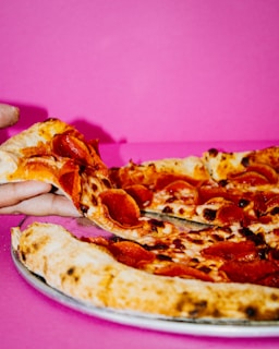 A pepperoni pizza is being served on a metal tray against a vibrant pink background. A hand is grabbing a slice, highlighting the melted cheese and crispy pepperoni.