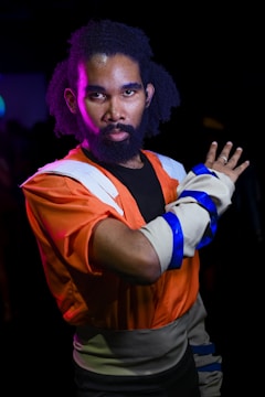 A person is wearing a costume inspired by an animated character, featuring an orange and blue outfit with white accents. The individual is posing with a serious expression and an outstretched hand. The background is dark, highlighting the vibrant colors of the costume.