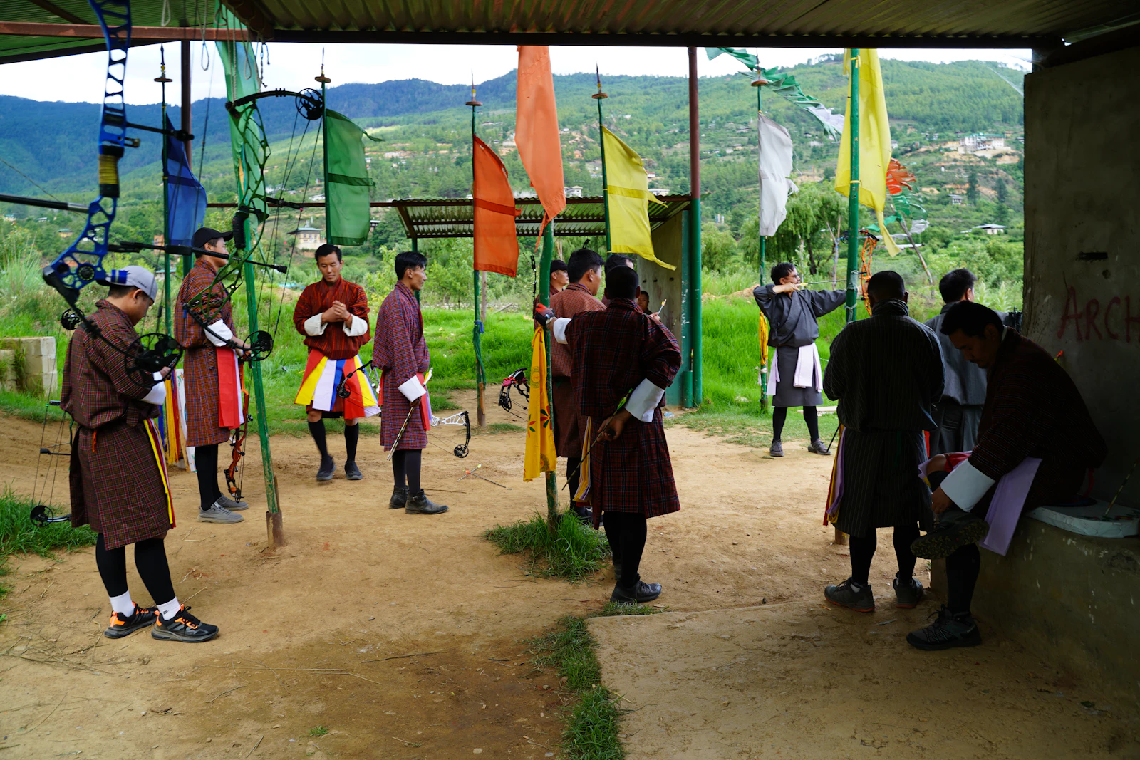 Archers in gho at a Bhutanese archery ground, bows and coloured flags along the line with green hills beyond