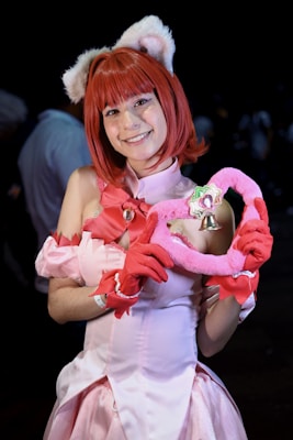 A person is dressed in a colorful, anime-inspired costume with cat ears and red hair, wearing a pink outfit with a detailed bow and holding a plush heart accessory.