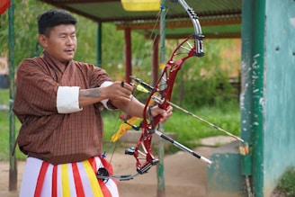 A man wearing traditional Bhutanese attire is actively engaged in archery. He is holding a modern, red compound bow with an arrow drawn, appearing focused and precise. The background shows a grassy area with a partially visible structure.