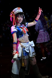 A person is dressed in a colorful, elaborate costume reminiscent of a sailor uniform, complete with a white hat adorned with a crest. The costume features blue, red, and white colors, with striped arm warmers and a decorative bow. Bubbles are floating around, creating a whimsical atmosphere.