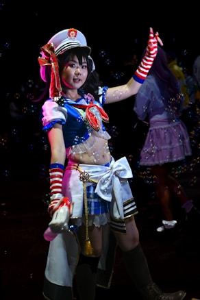 A person is dressed in a colorful, elaborate costume reminiscent of a sailor uniform, complete with a white hat adorned with a crest. The costume features blue, red, and white colors, with striped arm warmers and a decorative bow. Bubbles are floating around, creating a whimsical atmosphere.