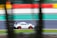 A white sports car with red accents and a large rear spoiler is speeding on a racetrack. The track features colorful barriers in orange, yellow, red, and green, with motion blur indicating high speed.