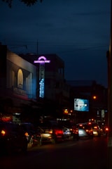 A nighttime urban scene with cars lined up on a busy street. Neon signs and billboards illuminate the buildings and the road. A particularly prominent sign advertises a karaoke venue. Distant traffic lights cast a red glow on the vehicles.