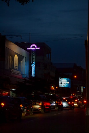 A nighttime urban scene with cars lined up on a busy street. Neon signs and billboards illuminate the buildings and the road. A particularly prominent sign advertises a karaoke venue. Distant traffic lights cast a red glow on the vehicles.