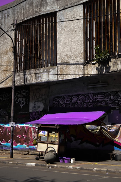 A street food cart with a vibrant purple canopy is positioned on a city street. The cart is bordered by a worn, graffiti-covered building featuring metal window bars. Brightly colored graffiti art decorates the wall behind the cart, adding an urban, artistic flair. Several utility poles and exposed wires are visible, contributing to the gritty urban atmosphere.
