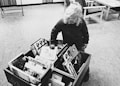 A young child with curly hair is standing and looking at a collection of books in a library or reading area. The books are situated in a wooden box on the ground, and the child appears to be focused and curious as they explore the titles.