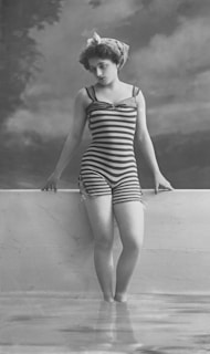 A vintage photograph features a woman standing in shallow water while leaning against a low wall. She is wearing a striped one-piece swimsuit and a headscarf, typical of early 20th-century fashion. The background is a painted backdrop of trees and sky, suggesting a studio setting.