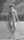 A vintage photograph features a woman standing in shallow water while leaning against a low wall. She is wearing a striped one-piece swimsuit and a headscarf, typical of early 20th-century fashion. The background is a painted backdrop of trees and sky, suggesting a studio setting.