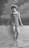 A vintage photograph features a woman standing in shallow water while leaning against a low wall. She is wearing a striped one-piece swimsuit and a headscarf, typical of early 20th-century fashion. The background is a painted backdrop of trees and sky, suggesting a studio setting.
