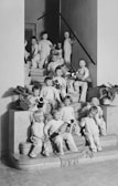 A group of young children, all dressed in similar light-colored outfits, are sitting and standing on a staircase. Some children are holding stuffed toys, and a few potted plants are placed on the steps. The setting appears to be indoors, with a simple, clean interior featuring a staircase with a railing and plain walls.
