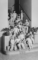 A group of young children, all dressed in similar light-colored outfits, are sitting and standing on a staircase. Some children are holding stuffed toys, and a few potted plants are placed on the steps. The setting appears to be indoors, with a simple, clean interior featuring a staircase with a railing and plain walls.