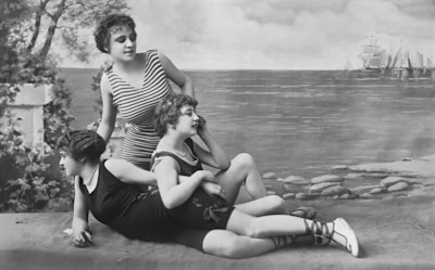 Three women in vintage swimsuits pose in front of a painted backdrop featuring the ocean and sailing ships. One is standing, while the other two are reclining beside her. The setting includes elements of nature like trees and rocks.