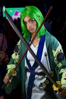A person is dressed in a cosplay outfit, featuring a vibrant green wig and a robe with intricate patterns. They are holding two swords in a crossed position, with a serious expression. The background is blurred, showcasing what appears to be a colorful display screen and a crowd.