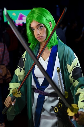 A person is dressed in a cosplay outfit, featuring a vibrant green wig and a robe with intricate patterns. They are holding two swords in a crossed position, with a serious expression. The background is blurred, showcasing what appears to be a colorful display screen and a crowd.