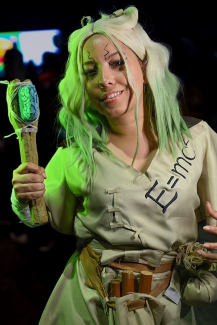 Person dressed in cosplay with light green hair, holding a staff with a green stone attached to it. They are wearing a beige outfit with a belt that has several items attached, including a few tubes. On their front, the equation E=mc^2 is written.