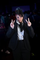 A person in a dark suit and white shirt stands in a crowded room, making peace signs with both hands. Their hair is styled simply with pink hair clips. The background is dimly lit with other people visible but out of focus.