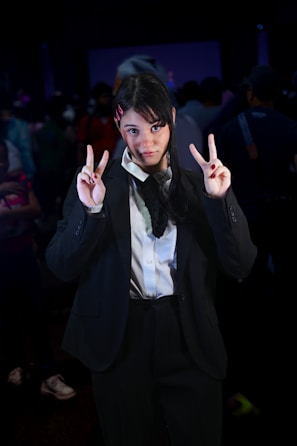 A person in a dark suit and white shirt stands in a crowded room, making peace signs with both hands. Their hair is styled simply with pink hair clips. The background is dimly lit with other people visible but out of focus.
