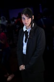 A person with long dark hair, wearing a black suit with a white shirt and black tie, is standing in a dimly lit environment. The individual appears to have a contemplative or slightly amused expression while looking towards the right. There are blurred figures in the background, suggesting a crowded setting.