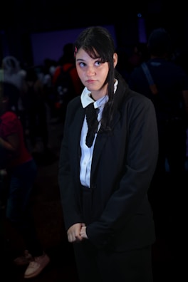 A person with long dark hair, wearing a black suit with a white shirt and black tie, is standing in a dimly lit environment. The individual appears to have a contemplative or slightly amused expression while looking towards the right. There are blurred figures in the background, suggesting a crowded setting.