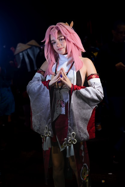 A person wearing an elaborate costume with pink hair styled in a long, flowing manner. The costume features detailed fabric with red, white, and gray colors, adorned with floral patterns and ornamental designs. The person is standing with hands posed in front of them, and there is a small badge or pass hanging from the neck.