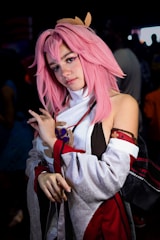 A person with pink hair is dressed in a detailed costume, potentially for cosplay. The costume includes a high-collared garment with intricate patterns, primarily in shades of white, black, and red. The individual is posing with a confident expression, in an environment with a dimly lit background that suggests a crowded setting.