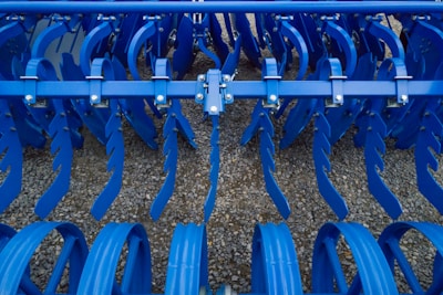 Industrial farming equipment with multiple rows of blue metal discs and blades arranged in a symmetrical pattern, against a gravel surface.