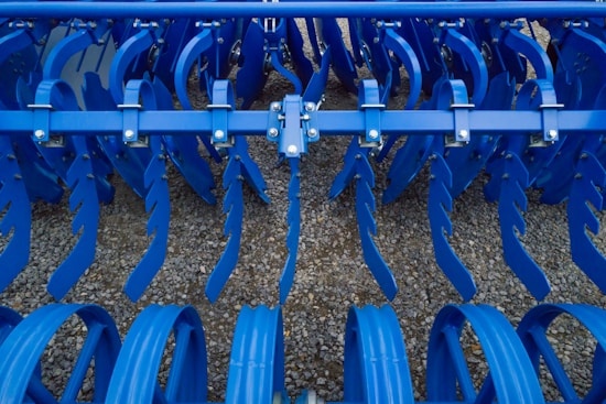 Industrial farming equipment with multiple rows of blue metal discs and blades arranged in a symmetrical pattern, against a gravel surface.