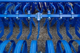 Industrial farming equipment with multiple rows of blue metal discs and blades arranged in a symmetrical pattern, against a gravel surface.