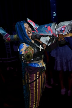 Person in a detailed, colorful cosplay costume holding a large, elaborately designed foam weapon. The costume features blue hair, intricate face paint, and vivid clothing with striped pants.