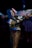 Person in a detailed, colorful cosplay costume holding a large, elaborately designed foam weapon. The costume features blue hair, intricate face paint, and vivid clothing with striped pants.