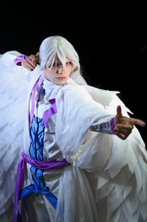 A person dressed in an elaborate costume with long white hair and a flowing white cape. The attire features blue and purple accents, suggesting a fantasy or anime-inspired theme. The person is striking a dynamic pose, pointing forward with a focused expression against a dark background.
