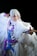 A person dressed in an elaborate costume with long white hair and a flowing white cape. The attire features blue and purple accents, suggesting a fantasy or anime-inspired theme. The person is striking a dynamic pose, pointing forward with a focused expression against a dark background.