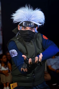 A person dressed in a detailed cosplay costume resembling a ninja character with spiky white hair, a metal headband, and a face mask. The individual makes a dynamic hand gesture, and the attire includes a dark green vest with distinct arm patches. The background is dark, with a few blurred people visible, emphasizing the character's dramatic pose and intricate costume.