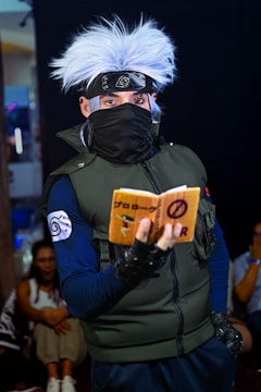 A person in elaborate costume with spiky white hair, a headband with a metallic emblem, a face mask, dark gloves, and a green vest is holding a small book with writing on it. The setting appears to be indoors with other people visible in the background.