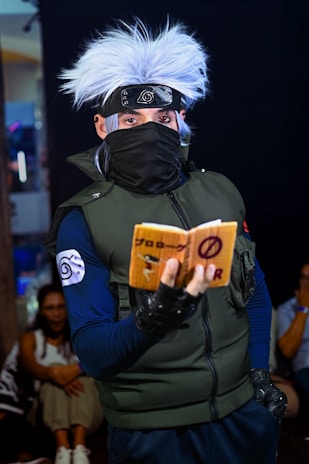 A person in elaborate costume with spiky white hair, a headband with a metallic emblem, a face mask, dark gloves, and a green vest is holding a small book with writing on it. The setting appears to be indoors with other people visible in the background.