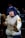 A person dressed in cosplay attire, featuring a white hoodie with a fur trim and a colorful patch on the sleeve, with blue hair and contact lenses. The pose suggests a martial arts stance, while the background includes several people in blurry focus, indicating a busy environment like a convention.
