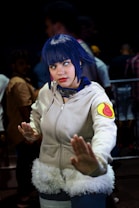 A person dressed in cosplay attire, featuring a white hoodie with a fur trim and a colorful patch on the sleeve, with blue hair and contact lenses. The pose suggests a martial arts stance, while the background includes several people in blurry focus, indicating a busy environment like a convention.