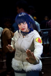A person dressed in cosplay attire, featuring a white hoodie with a fur trim and a colorful patch on the sleeve, with blue hair and contact lenses. The pose suggests a martial arts stance, while the background includes several people in blurry focus, indicating a busy environment like a convention.