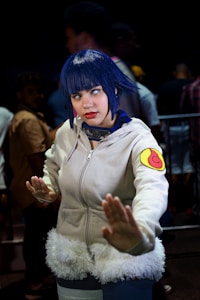 A person dressed in cosplay attire, featuring a white hoodie with a fur trim and a colorful patch on the sleeve, with blue hair and contact lenses. The pose suggests a martial arts stance, while the background includes several people in blurry focus, indicating a busy environment like a convention.