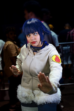 A person dressed in cosplay attire, featuring a white hoodie with a fur trim and a colorful patch on the sleeve, with blue hair and contact lenses. The pose suggests a martial arts stance, while the background includes several people in blurry focus, indicating a busy environment like a convention.